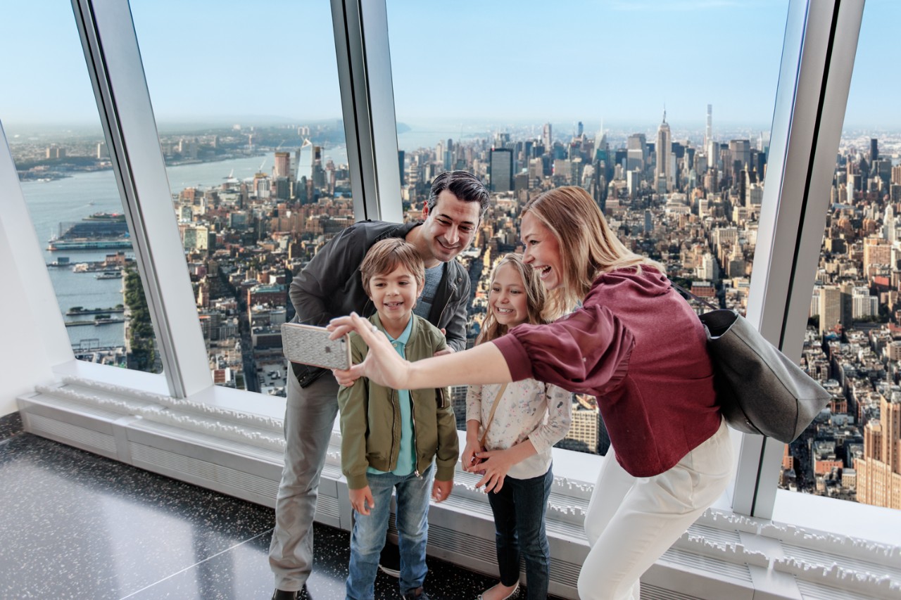 A family takes a selfie at One World Observatory with a view of the New York City skyline behind them